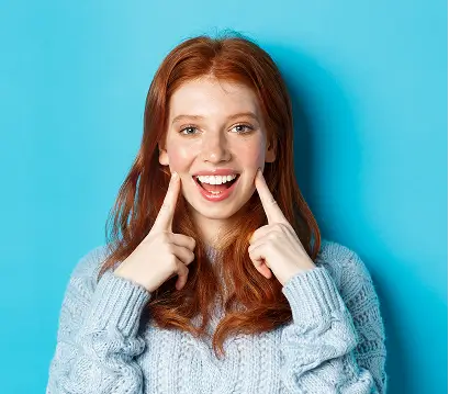 Joven pelirroja sonriendo y señalando sus mejillas alegremente.